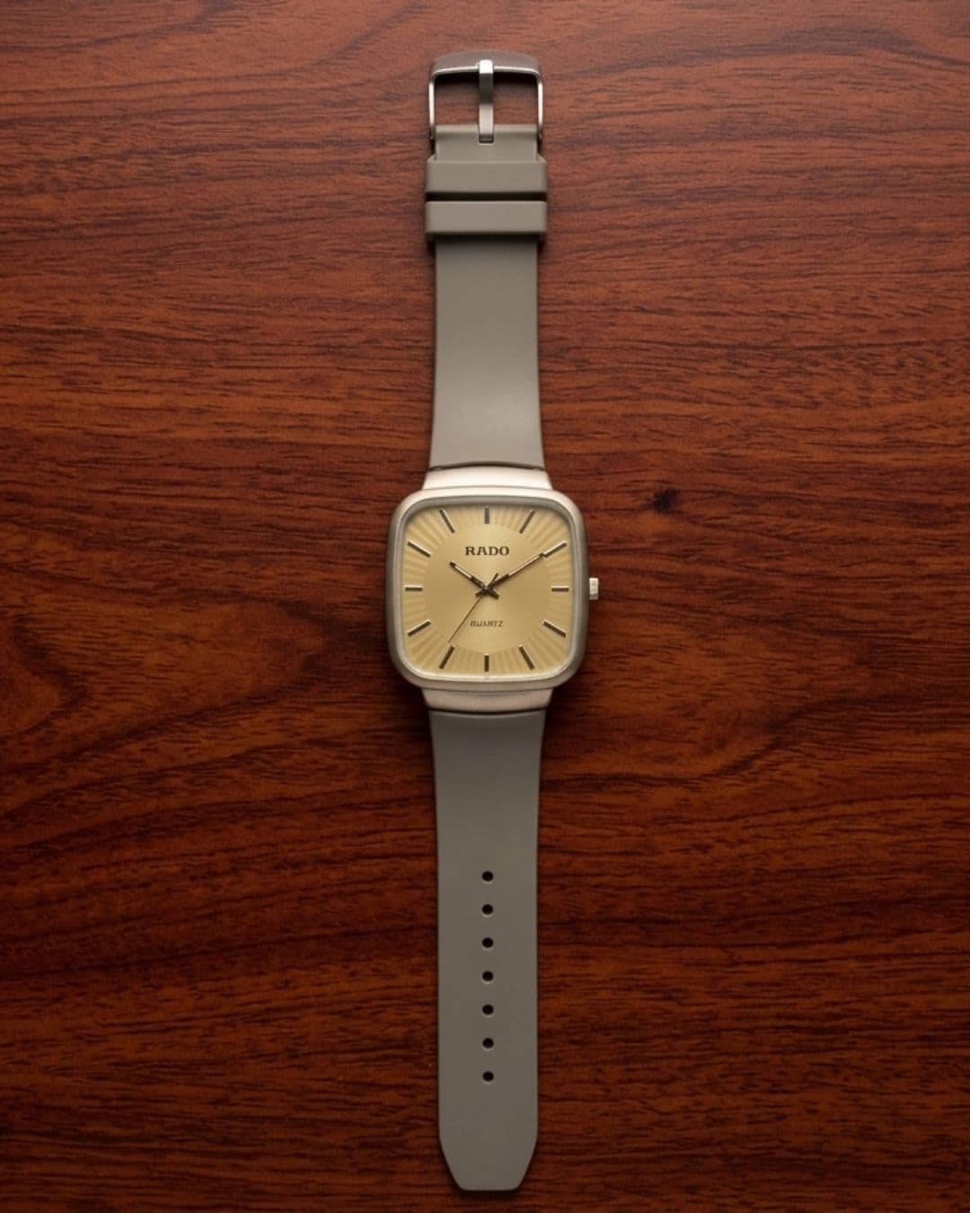 A Rado watch with a square beige dial and a light gray strap, laid flat on a dark brown wooden surface.